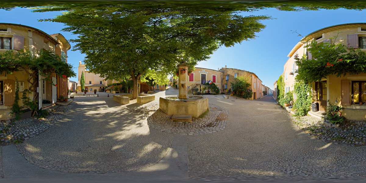 Equirectangular 360° view of a Provençal village square in the morning, with a stone fountain in the centre under a large tree, traditional houses with shuttered windows, and the Mairie building flying the French and European flags