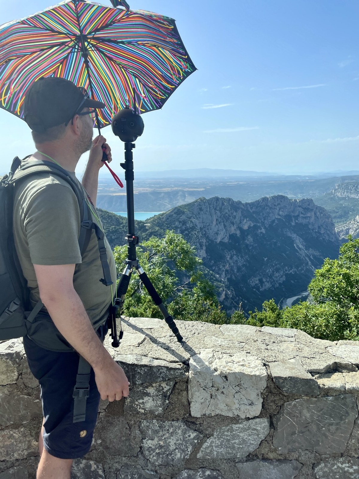 Eric standing at a stone viewpoint above the Verdon Gorge holding a colourful umbrella over a 360° camera mounted on a monopod, deep canyon and lake visible in the distance