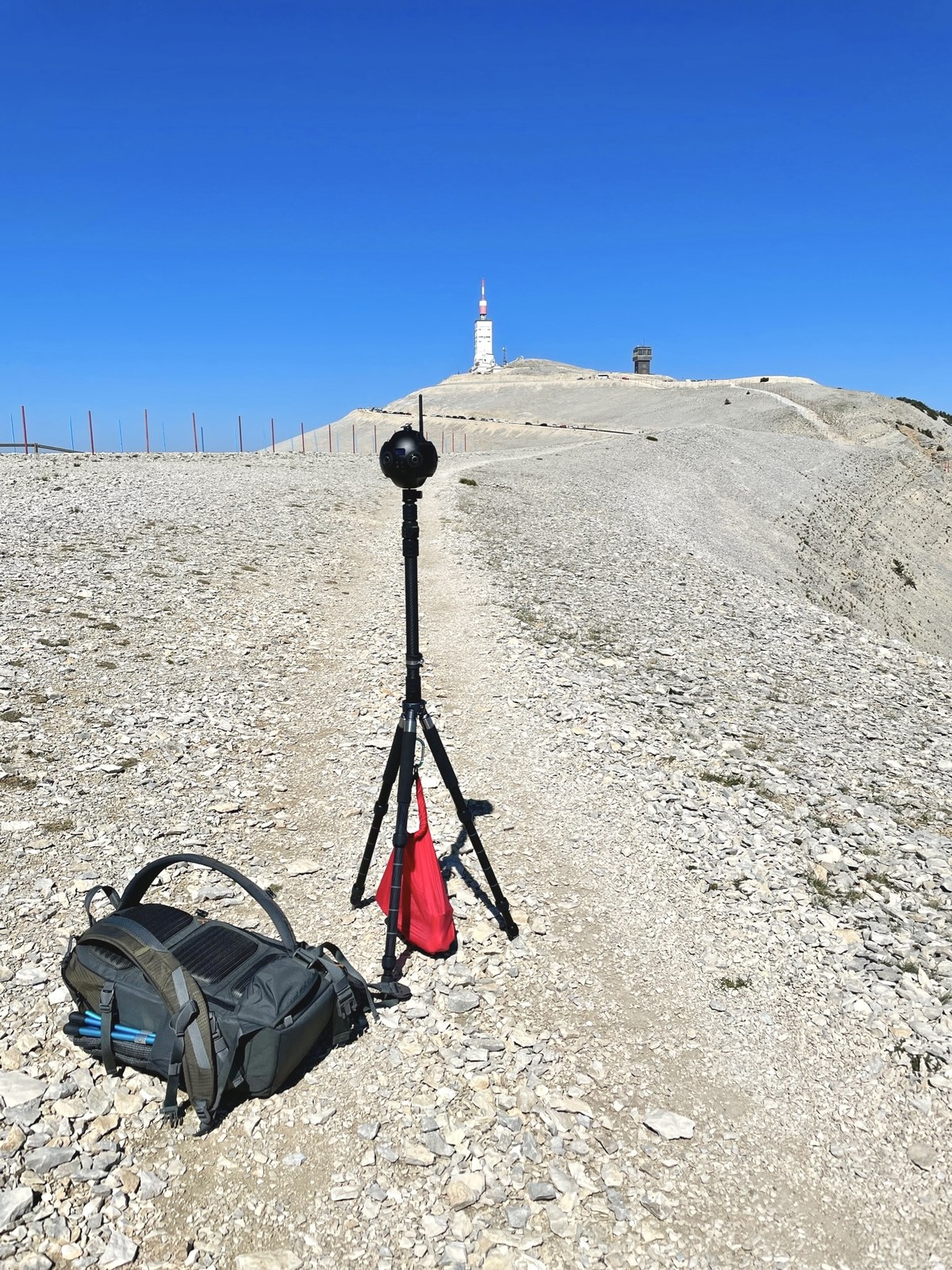 A 360° camera on a tall tripod stands on the white limestone summit ridge of Mont Ventoux, with the iconic weather station and tower visible in the distance under a deep blue sky