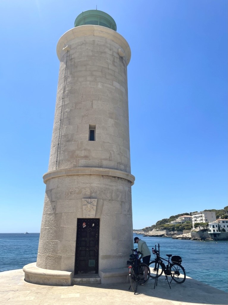A small stone lighthouse at the end of a harbour breakwater under bright blue sky, with two bicycles parked at its base, Mediterranean Sea and coastal village visible in the background