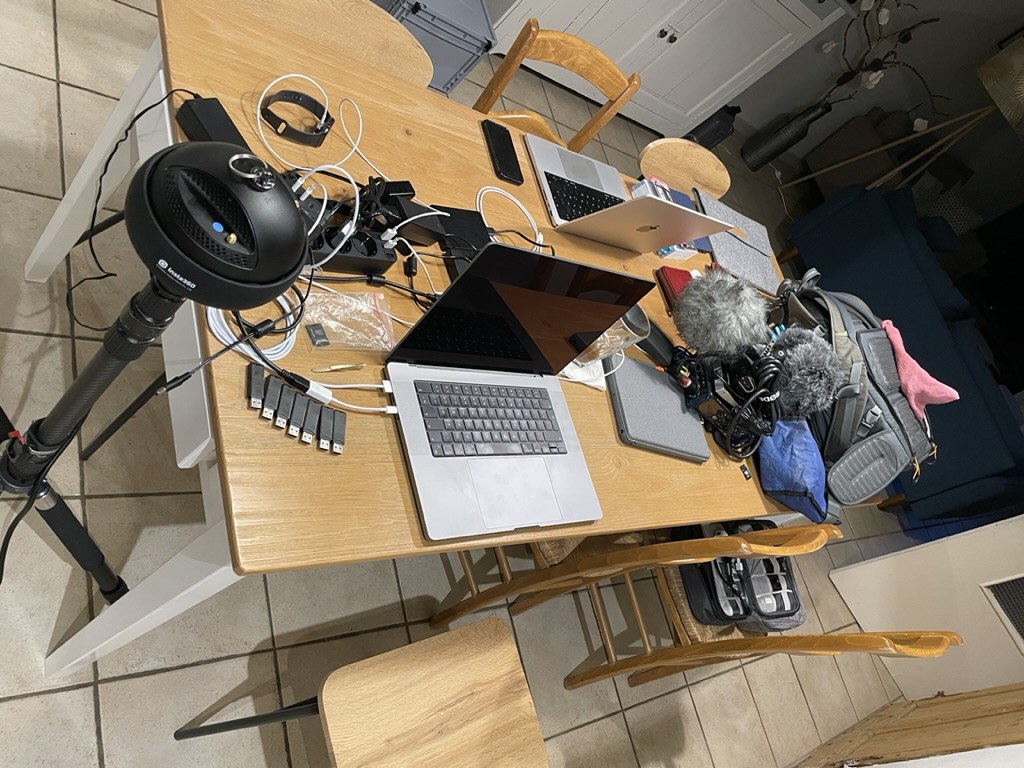 Overhead view of a wooden dining table workstation showing two MacBook laptops, the 360° camera on its stand, microphone with windshield, multiple SD card readers in a row, and other production equipment